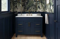 A trendy navy blue bathroom with a furniture style blue vanity with a solid white countertop, gold/brushed brass hardware, blue wainscot panels, and a fashionable blue, green, and white wallpaper.