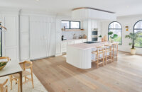 A bright, all-white kitchen with an elegant flat panel cabinet door style and unique reeded details with a curved kitchen island and dropdown ceiling feature.