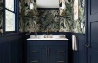A trendy navy blue bathroom with a furniture style blue vanity with a solid white countertop, gold/brushed brass hardware, blue wainscot panels, and a fashionable blue, green, and white wallpaper.