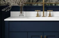 A trendy navy blue bathroom with a furniture style blue vanity with a solid white countertop, gold/brushed brass hardware, blue wainscot panels, and a fashionable blue, green, and white wallpaper.