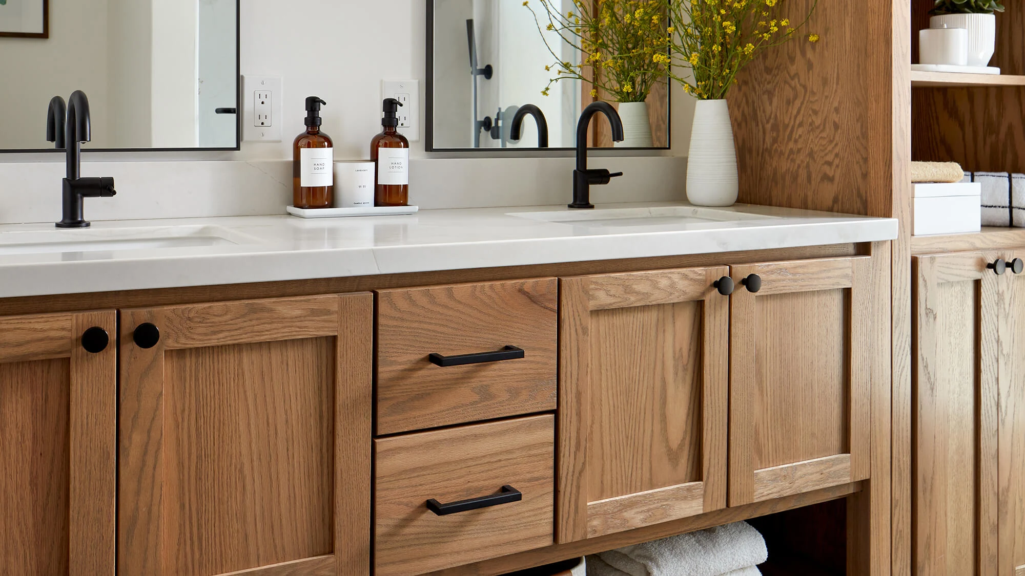 A close up angle of a warm oak vanity with two sinks with black faucets and a crisp, white countertop.