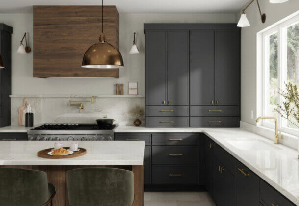 Modern kitchen with gray cabinets, brass hardware, white countertops, a wooden island with two green chairs, pendant lights, a stove, and a large window with natural light and decorative plants.