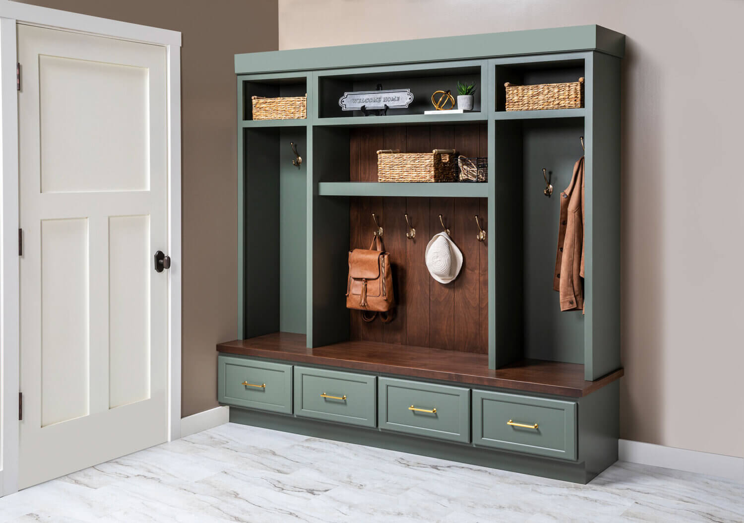 Custom mudroom built-in with sage-green Grove cabinetry, dark Kodiak-stained Alder wood bench and backing, woven storage baskets, brass hooks with hanging bags and hat, and four lower drawers with brass pulls