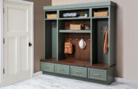 Custom mudroom built-in with sage-green Grove cabinetry, dark Kodiak-stained Alder wood bench and backing, woven storage baskets, brass hooks with hanging bags and hat, and four lower drawers with brass pulls