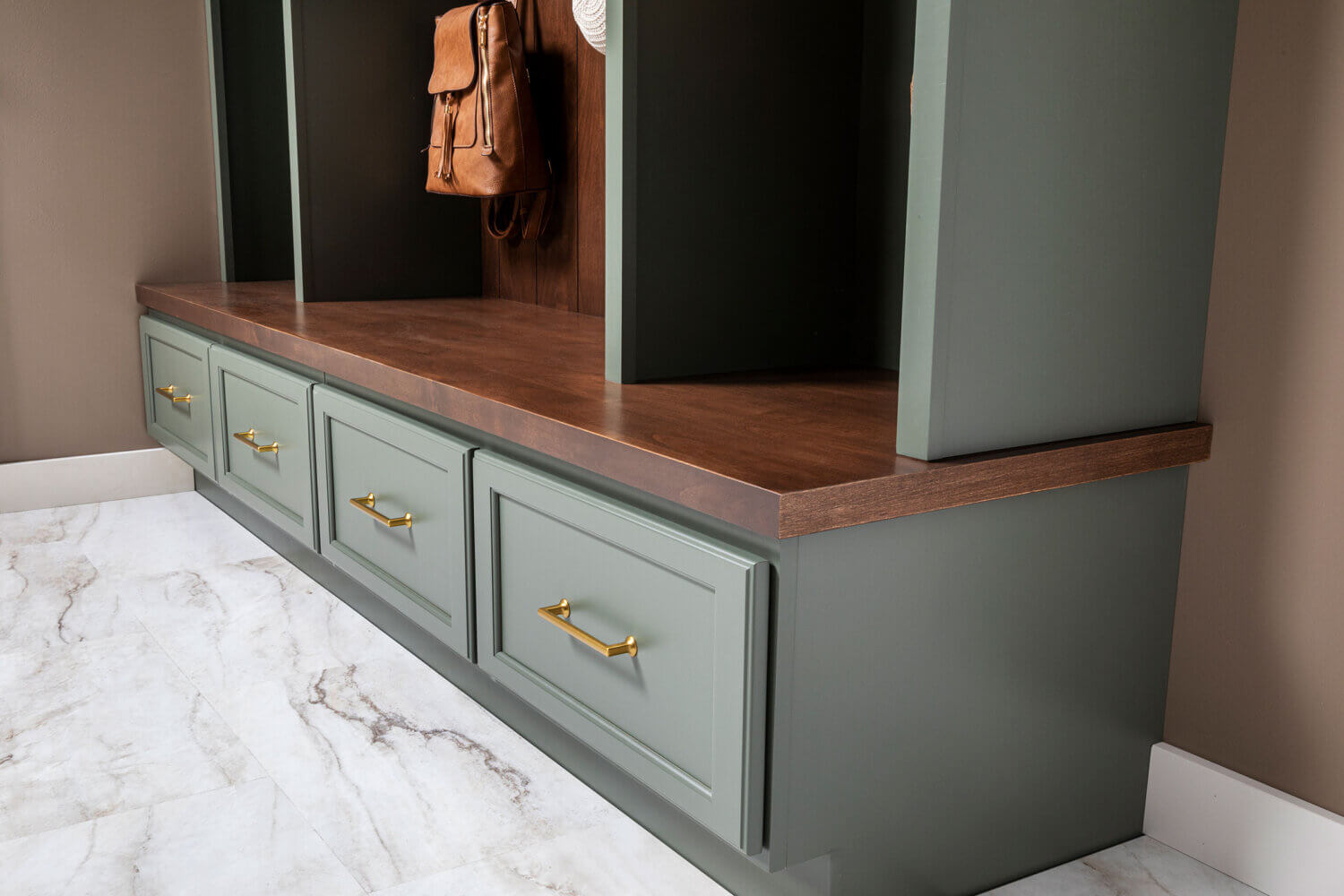 Custom mudroom built-in with sage-green Grove cabinetry, dark Kodiak-stained Alder wood bench and backing, woven storage baskets, brass hooks with hanging bags and hat, and four lower drawers with brass pulls