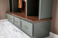 Custom mudroom built-in with sage-green Grove cabinetry, dark Kodiak-stained Alder wood bench and backing, woven storage baskets, brass hooks with hanging bags and hat, and four lower drawers with brass pulls