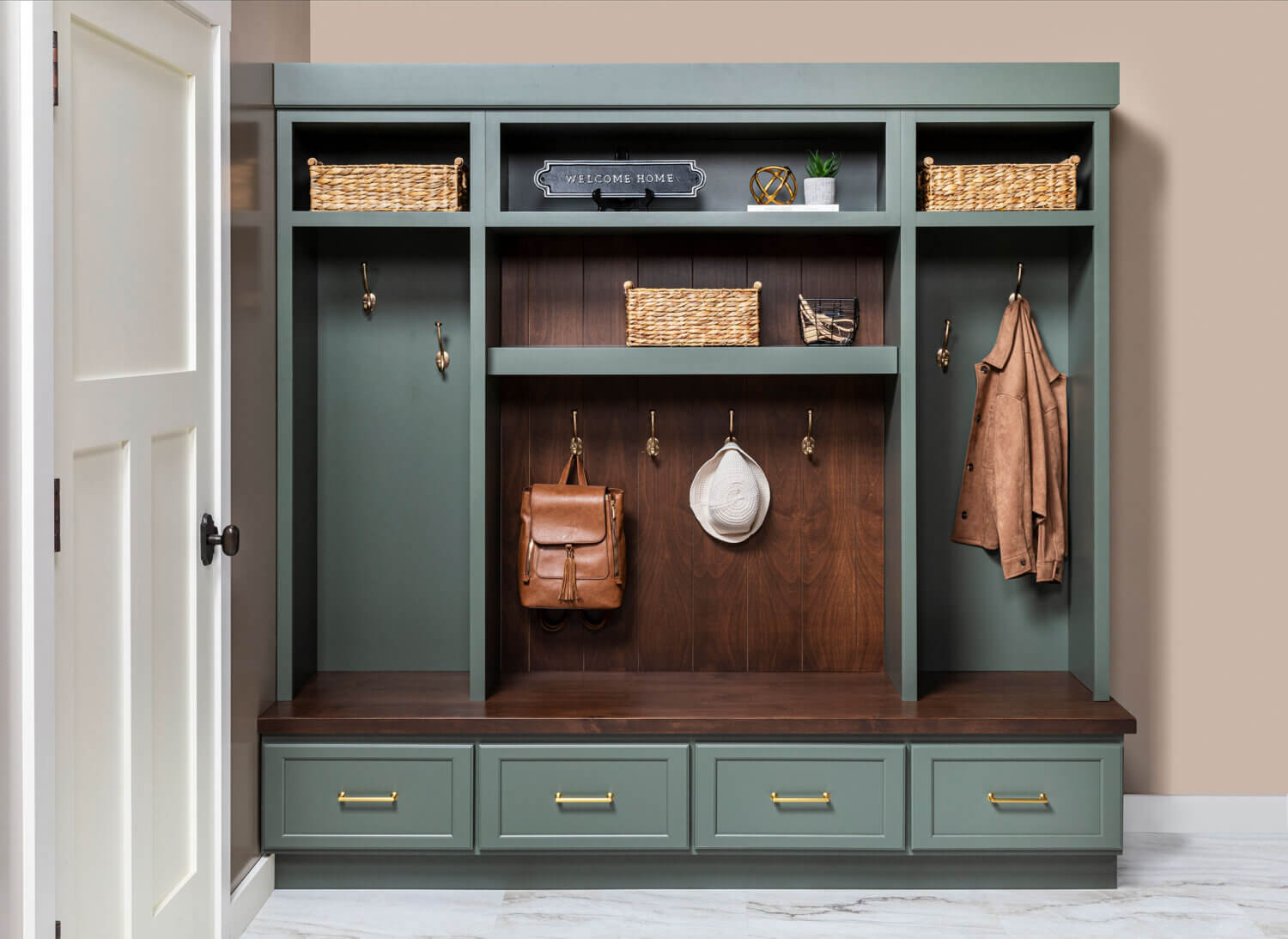 Custom mudroom built-in with sage-green Grove cabinetry, dark Kodiak-stained Alder wood bench and backing, woven storage baskets, brass hooks with hanging bags and hat, and four lower drawers with brass pulls