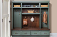 Custom mudroom built-in with sage-green Grove cabinetry, dark Kodiak-stained Alder wood bench and backing, woven storage baskets, brass hooks with hanging bags and hat, and four lower drawers with brass pulls